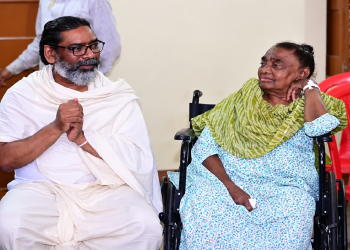 रूपी सोरेन व्हीलचेयर पर बैठी हैं और उनके पास हेमंत सोरेन सफेद वस्त्रों में बैठे नजर आ रहे हैं।
