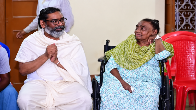 रूपी सोरेन व्हीलचेयर पर बैठी हैं और उनके पास हेमंत सोरेन सफेद वस्त्रों में बैठे नजर आ रहे हैं।